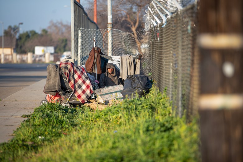 A person sits in a makeshift tent along a barbed wire fence near Highway 99 in southwest Fresno on Feb. 11, 2022. The fence blocks out a grass area that used to be a homeless encampment. Photo by Larry Valenzuela for CalMatters/CatchLight Local