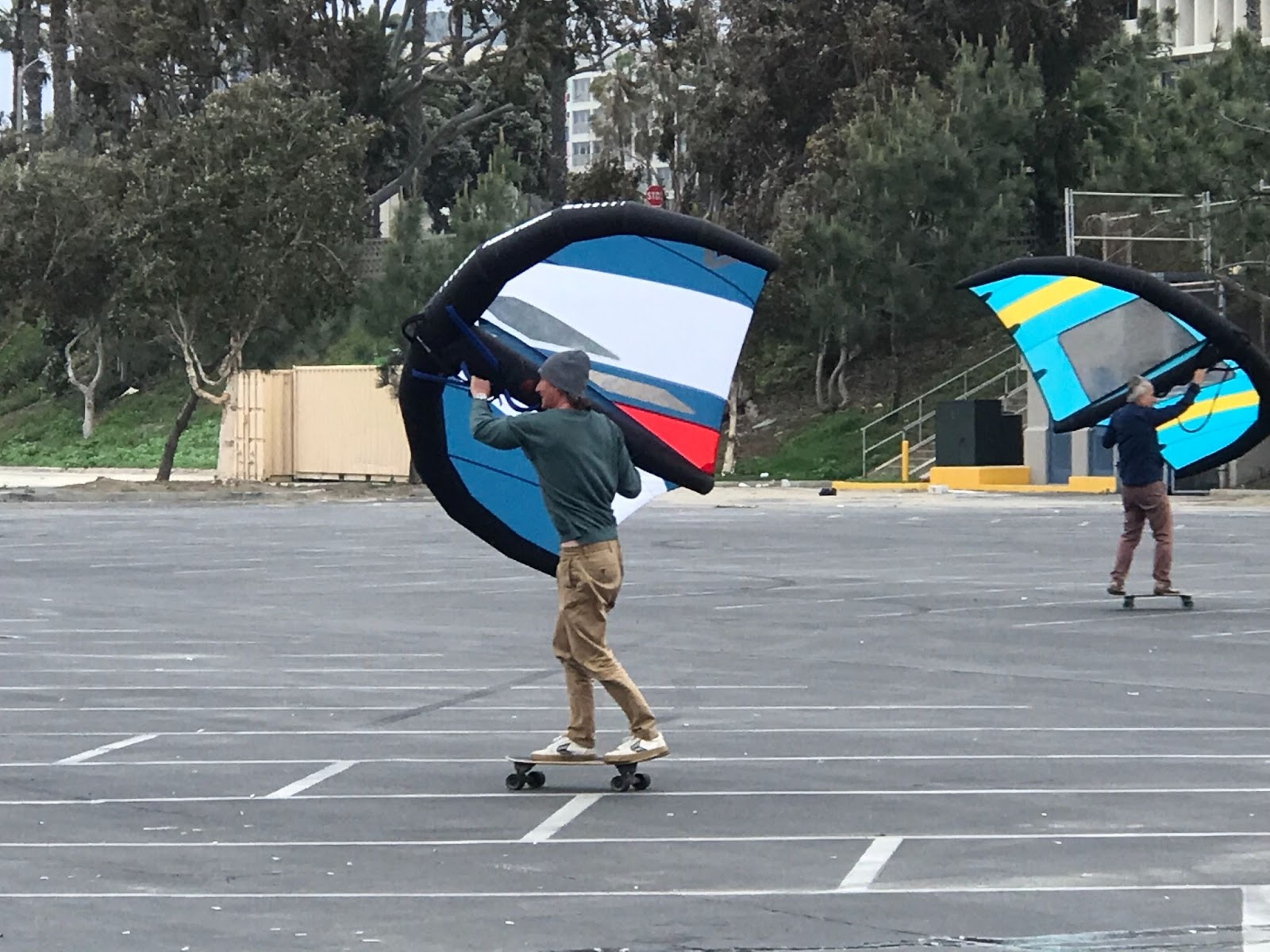 Two people using kites to catch wind as they ride skateboards in a parking lot