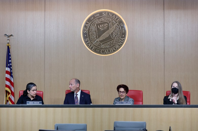 Lawmakers sit in read chairs on a long and elevated desk in a hearing room at the state capitol. A seal of the Senate of the State of California rest on the wall behind them.