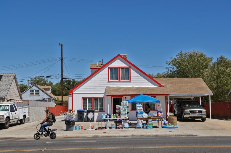 A street vendor sells fans, mini pools and other products outside a white and red two-story house as a man in a bicycle passes by.