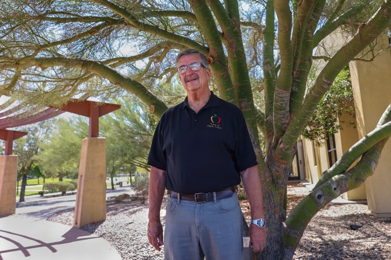 A person wearing glasses, a black polo shirt and grey pants stands under a palo verde tree outside of a building during a sunny day.