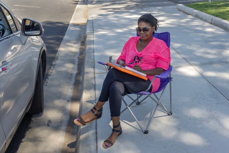 A person wearing a pink shirt, black pants and sandals sits in a folding chair on a sidewalk while writing in a notebook.