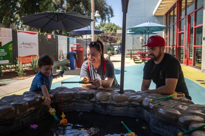 A child, on the left side of the frame, places a toy fishing hook in to a small water well with other toys floating around, as his mother and father play next to him.