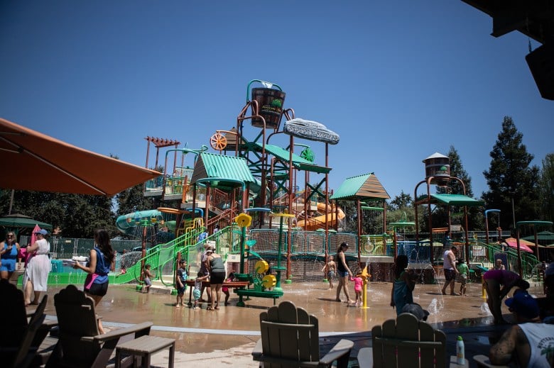 A wide view of people at a water park with various slides, water toys and splash pads.