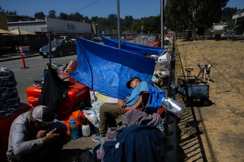 An unhoused man sits slumped on a chair next to a blue tarp and surrounded by possesion near him. Another unhoused man sits nearby him at a camp outside the Housing Matters shelter in Santa Cruz on Aug. 7, 2024. Photo by Manuel Orbegozo for CalMatters