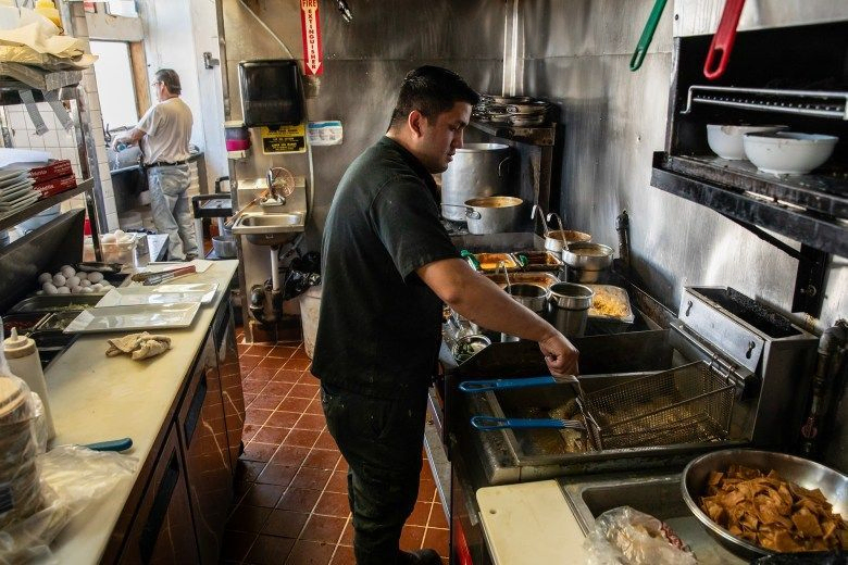 A cook dressed in a black outfit works in the kitchen of a restaurant, at the fryer. Another person, in the background, wearing denim and a white shirt, washes dishes.