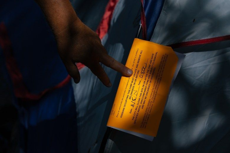 An unhoused person touches an orange eviction notice on a tent set up on Pacific Avenue in Santa Cruz on Aug. 7, 2024. Photo by Manuel Orbegozo for CalMatter