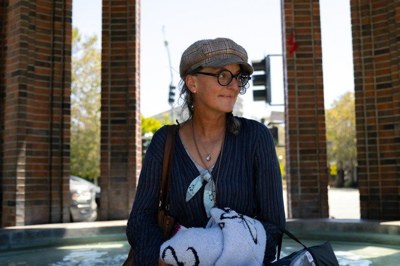 Stephanie Ross, 49, wearing a plaid hat and holding a piece of clothing and two bags, looks to her left in Santa Cruz on Aug. 7, 2024. Photo by Manuel Orbegozo for CalMatters