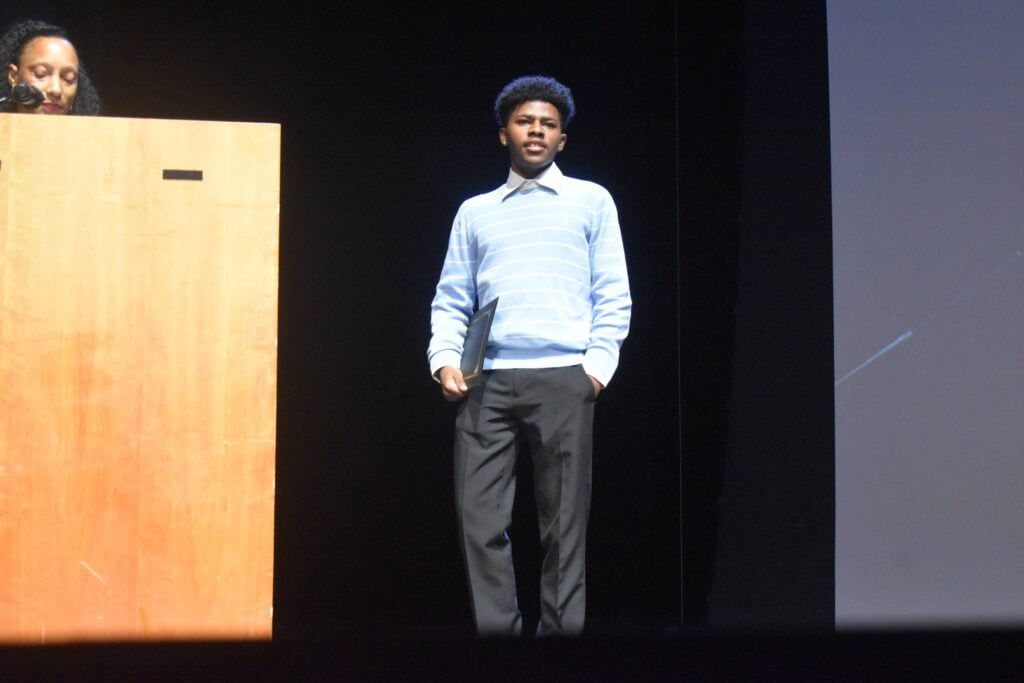 A Black male teenager receives an educational award on stage.