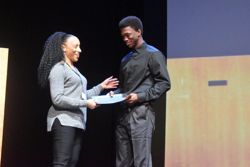 A Black male teenager receives an educational award for a project.