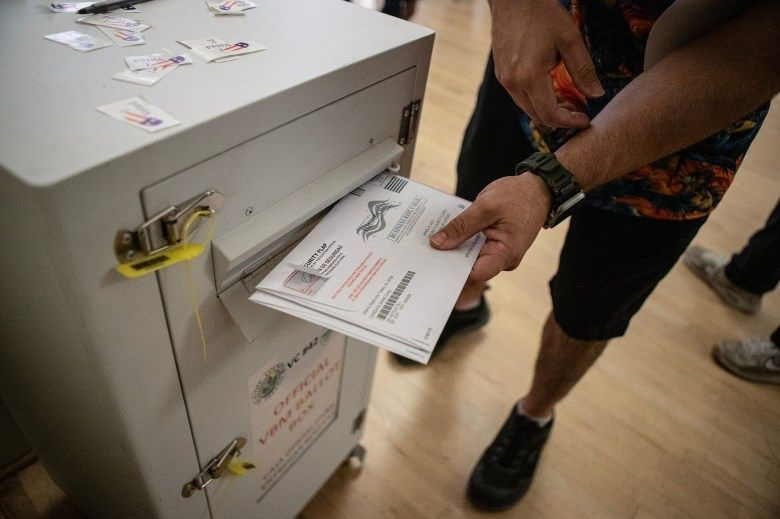 A person deposits completed ballots into an official, secured ballot drop box. The drop box is white with labels and secured with yellow zip ties. 