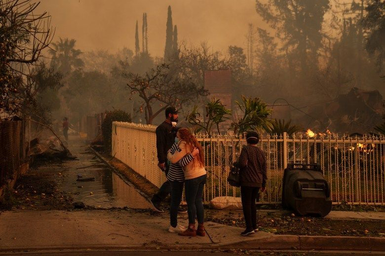 Two people hug a burned-down structure behind a fence while other people walk around in the destroyed neighborhood. Smoke lingers in the background, and green and palm trees remain in the haze.