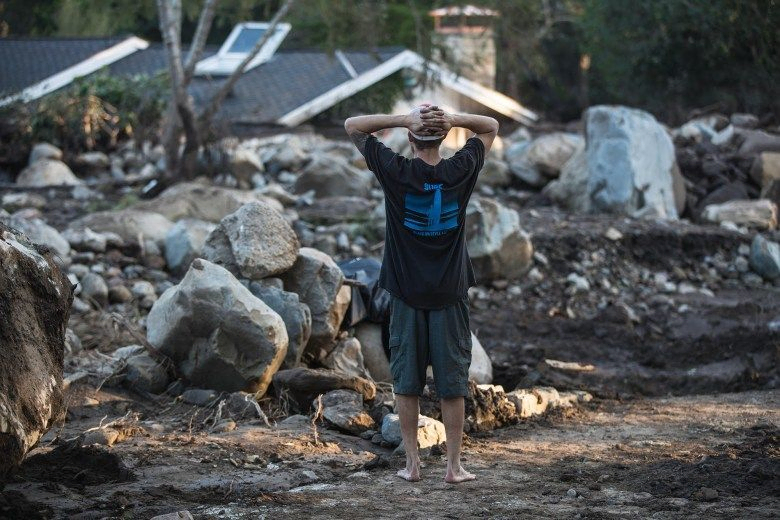 The back of a person with their hands resting on the back of their head as they look at the wreckage of a home in front of them amidst rocks and mud.