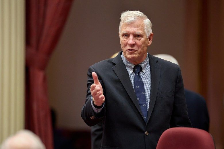 A lawmaker, with white hair and wearing a black suit with a blue tie and blue patterned-shirt, gestures with their right hand as they speak in front of other lawmakers.